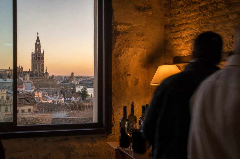 Seville rooftops at golden hour with the cathedral bell tower rising above tiled roofs and the Guadalquivir visible in the distance