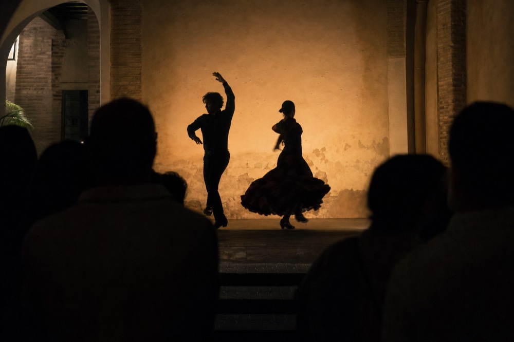 Two dancers in silhouette against a warm-lit back wall, one arm raised, skirt caught mid-turn, the audience visible only as dark shapes