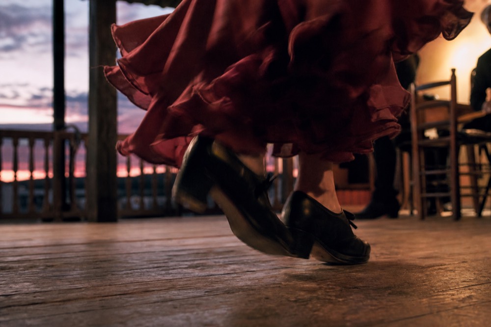 Close view of a dancer's shoes striking a worn wooden tablao stage, motion blur on the heel, ruffled hem lifted just above the boards
