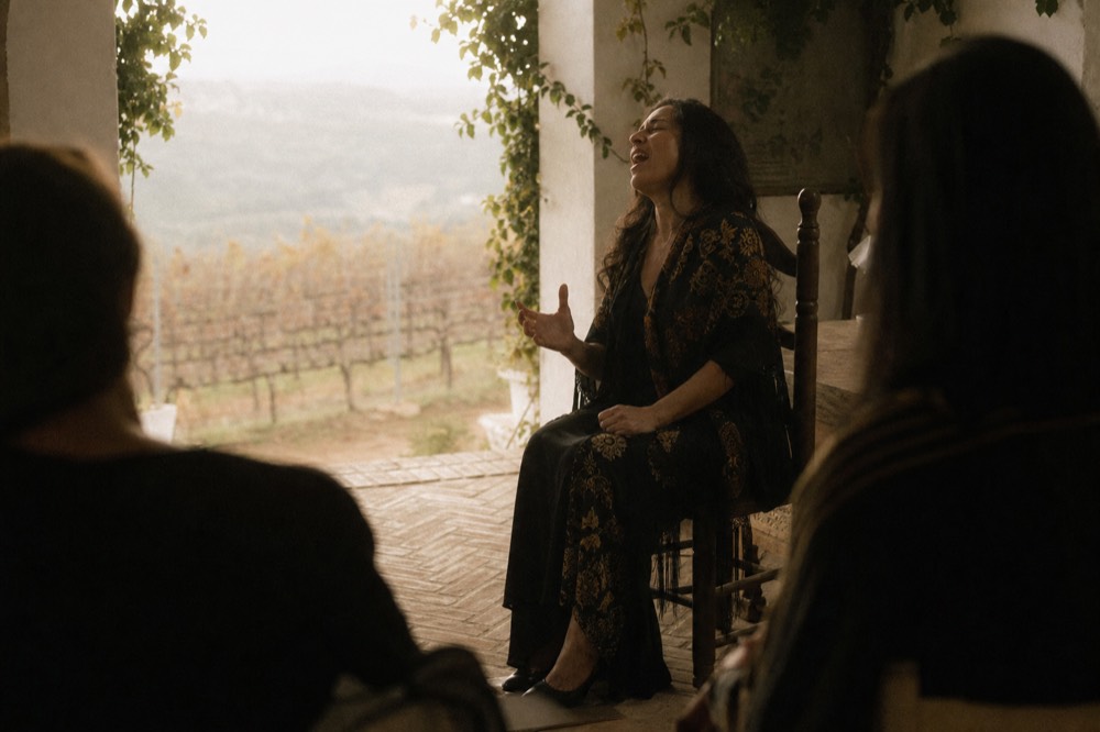 Flamenco singer seated on a wooden chair mid-phrase, head tilted back, hand raised, no microphone stand anywhere in the frame
