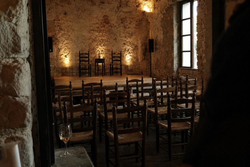 Rows of empty wooden chairs facing a small raised plank stage before a show, a single glass of sherry on a side ledge