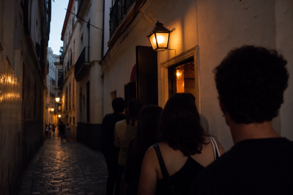 Narrow Seville street at dusk with a small illuminated venue doorway, a short queue of people waiting under a lantern
