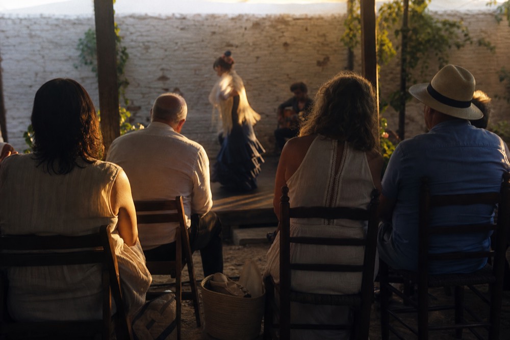 Front row of a small tablao seen from behind, seated guests in light summer clothes leaning forward, bags tucked under the chairs