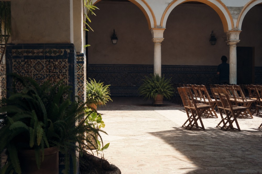 Sunlit Seville courtyard with azulejo tiles, potted ferns and a shaded arcade of arches where rows of chairs are set out