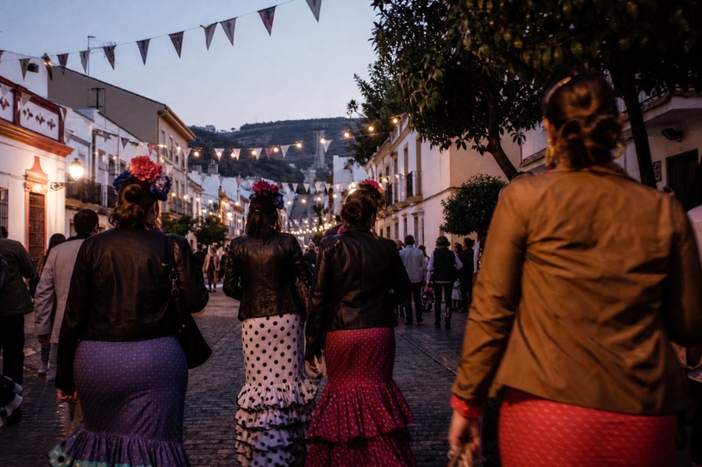 Crowded Seville street during a spring festival, ruffled polka-dot skirts and dark jackets seen from behind, bunting strung overhead