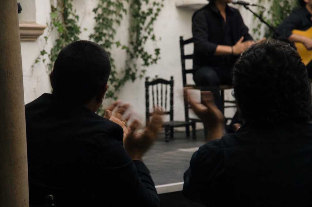 Two seated palmeros clapping in rhythm at the edge of a flamenco stage, hands blurred, singer beside them out of focus