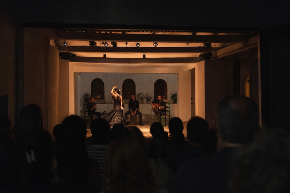 Darkened flamenco room with the stage brightly lit and the audience in shadow, no screens raised, every face turned forward