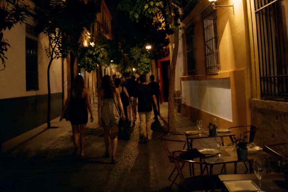 Lamplit Seville street after dark with people walking towards a small venue doorway, empty restaurant terrace tables set for later